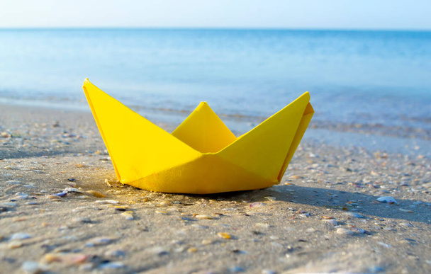 Small paper yellow boat on sand near water on background of sea waves - Foto, Imagem