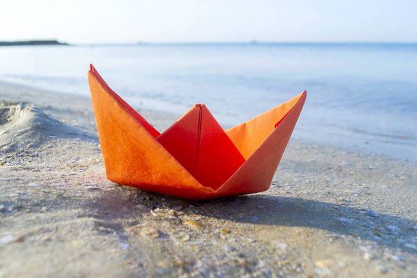 Невеликий паперовий помаранчевий човен на піску біля води на фоні морських хвиль
 - Фото, зображення
