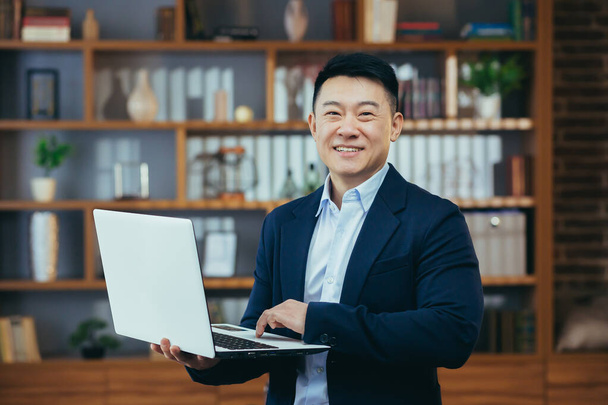 Homme d'affaires heureux et prospère debout travaillant sur un ordinateur portable, souriant et regardant la caméra, asiatique travaillant dans un bureau à domicile classique pour le dîner - Photo, image