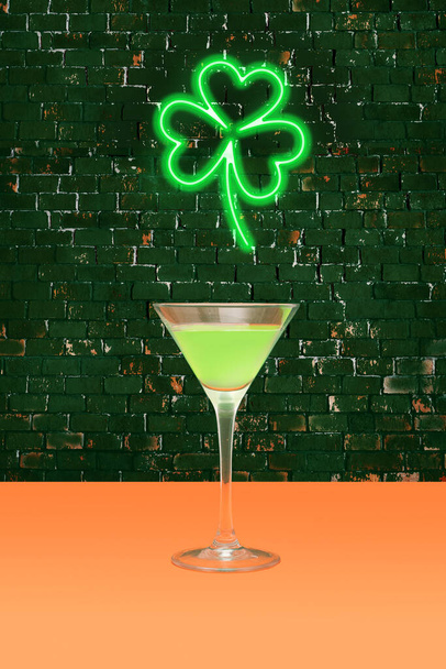 Lucky pub minimal concept. Martini glass with green cocktail on terracotta table. Shamrock green neon sign above on dark brick background. - Photo, Image