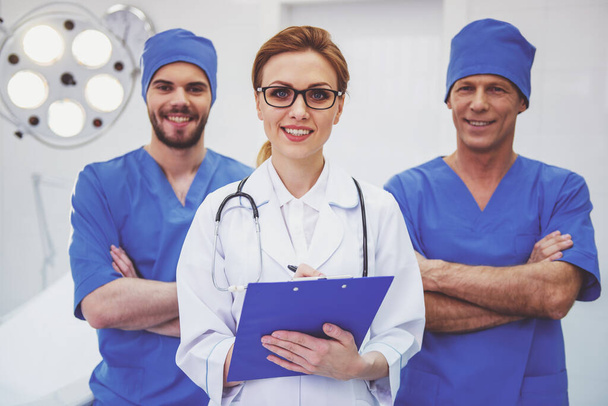 Belle femme médecin au premier plan et deux hommes regardent la caméra et sourient, debout dans une salle d'opération moderne
 - Photo, image
