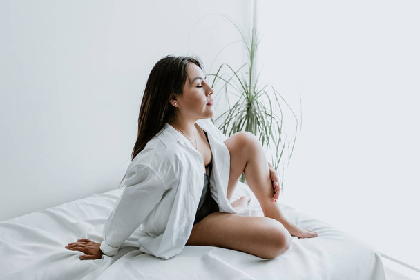 Young Latin woman sitting on bed, wearing white robe, showing legs at home in Mexico city - Foto, Bild