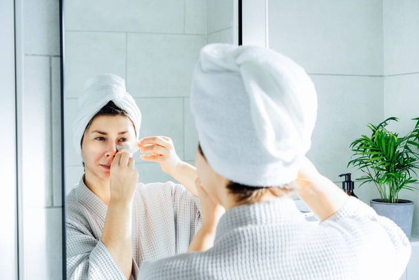 Woman in a bathrobe with a towel on her head applying patches under eye area, looking at mirror, enjoying daily antiwrinkle antiage routine in bathroom after morning shower. Home Beauty self-care - Photo, image