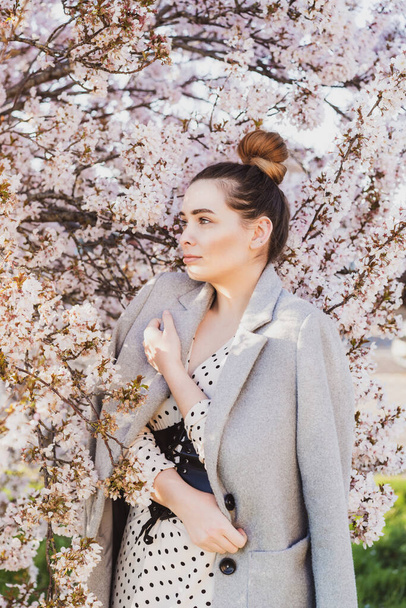 Spring Beautiful romantic woman standing in blooming garden. Tenderness Young model in a gray coat. Vertical card. Sensitive mood. Selective focus, coy space. - Foto, Imagen