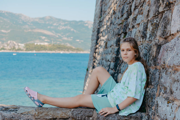 Beautiful girl sitting on a stone wall, in background is the blue sea, Budva, Montenegro. - Photo, Image