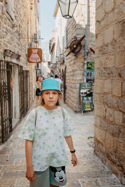 Girl Tourist Walking Through Ancient Narrow Street On A Beautiful Summer Day In Mediterranean Medieval City, Old Town Budva, Montenegro. Young Beautiful Cheerful Woman Walking On Old Street At - Foto, afbeelding