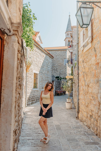 Girl Tourist Walking Through Ancient Narrow Street On A Beautiful Summer Day In MEDITERRANEAN MEDIEVAL CITY, OLD TOWN BUDVA, MONTENEGRO. Young Beautiful Cheerful Woman Walking On Old Street At - Foto, immagini
