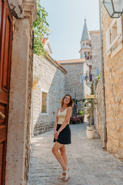 Girl Tourist Walking Through Ancient Narrow Street On A Beautiful Summer Day In MEDITERRANEAN MEDIEVAL CITY, OLD TOWN BUDVA, MONTENEGRO. Young Beautiful Cheerful Woman Walking On Old Street At - Zdjęcie, obraz