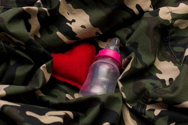 Askeri üniformada kırmızı kalp ve bebek şişesi. Aşk ve savaş kavramı - Fotoğraf, Görsel