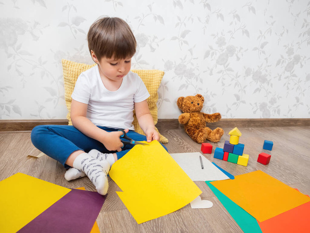 Toddler boy learns to cut colored paper with scissors. Kid sits on floor in kids room with toy blocks and teddy bear. Educational classes for children. Developing feeling sensations and fine motor skills at home. - Foto, afbeelding