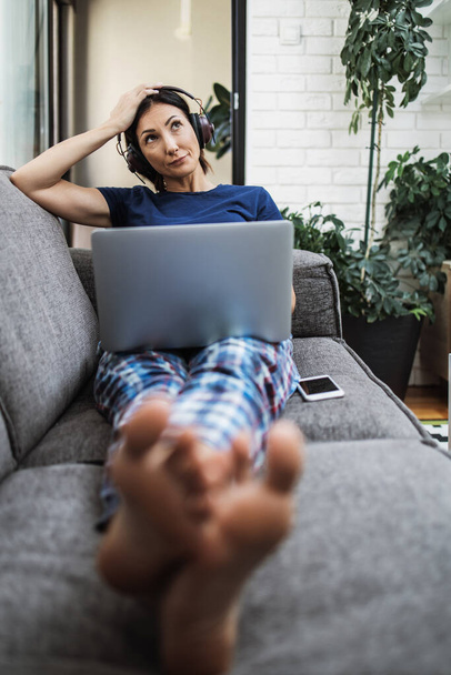 Young adult woman lying on sofa in her living room and using laptop computer for web surfing and social media communication and chat. - Foto, immagini