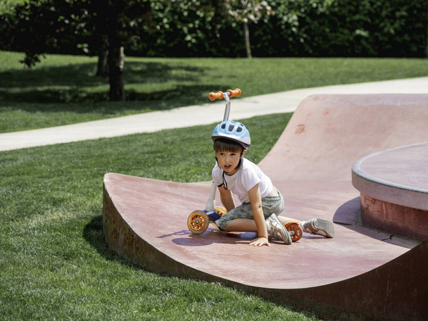 Little boy fell off kick scooter while riding in skate park. Special concrete bowl structures in urban park. Training to skate at summer. - 写真・画像