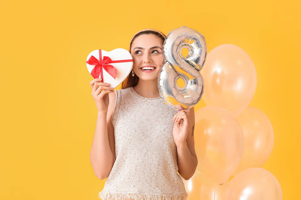 Счастливая молодая женщина с подарком и воздушными шарами на желтом фоне. Международный женский день - Фото, изображение