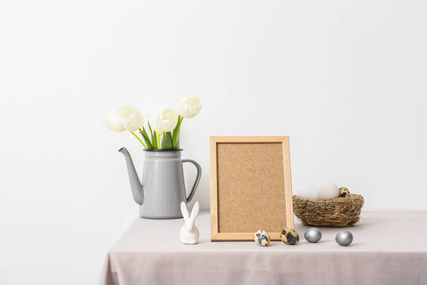 Blank photo frame, nest with Easter eggs, tulips and rabbit on table near light wall - Photo, Image