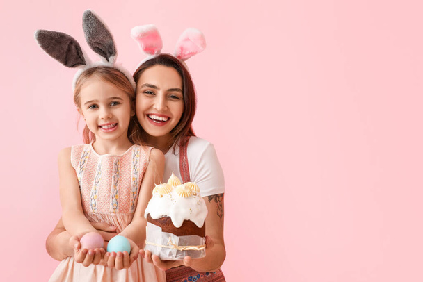Little girl, her mother with tasty Easter cake and eggs on pink background - Fotó, kép