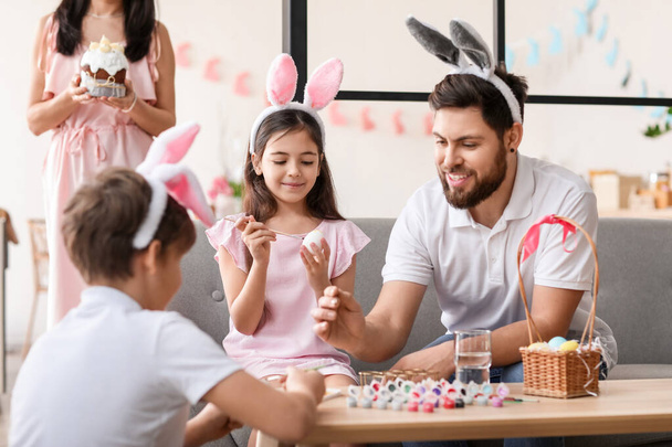 Маленька дівчинка в кролячих вухах зі своєю сім'єю малює великодні яйця вдома
 - Фото, зображення