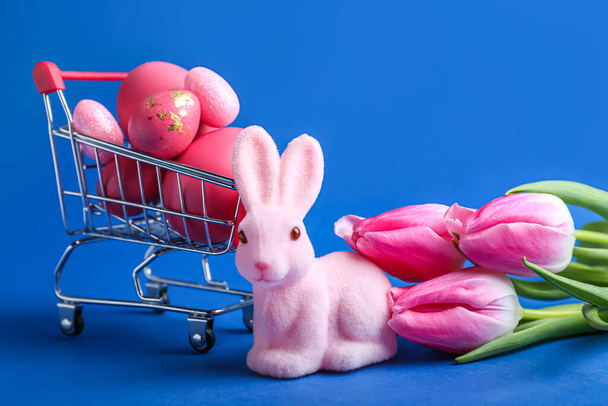 Cute Easter bunny, shopping trolley with painted eggs and flowers on blue background - Foto, Imagem