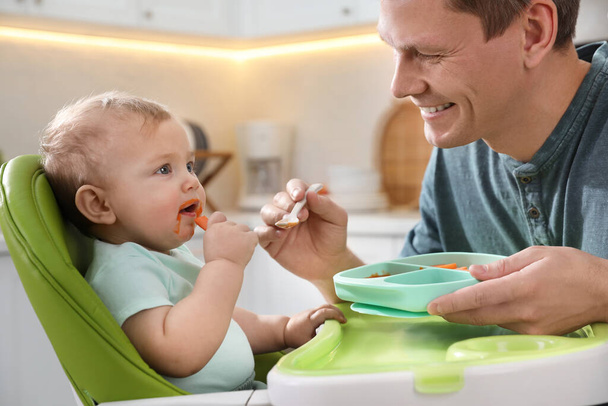 Father feeding his cute little baby in kitchen - Фото, изображение