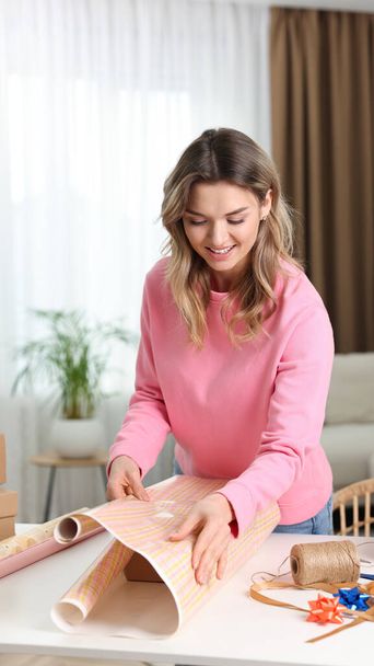 Beautiful young woman wrapping gift at table in living room - Photo, Image