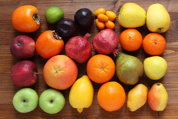 茶色の木製のテーブルの上の果物の異なる種類。上からの眺め。閉鎖だ。柿、リンゴ、プラム、ザクロ、梨、オレンジ、みかん、ベルガモット、クワット、ライム、グレープフルーツ. - 写真・画像