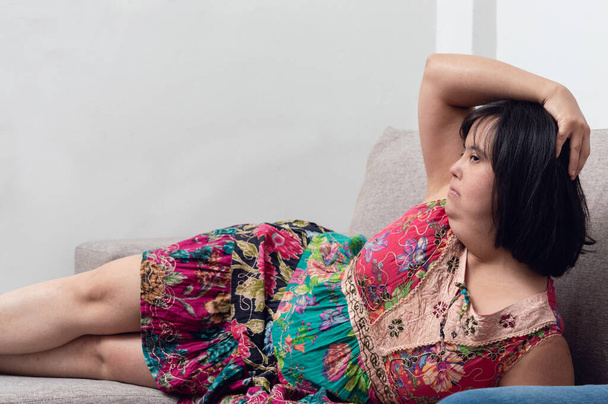 schöne kaukasische lateinamerikanische junge Frau mit Down-Syndrom liegend auf dem Sofa im Wohnzimmer posiert, während sie fernsieht, Inklusion und weibliches Machtkonzept. - Foto, Bild