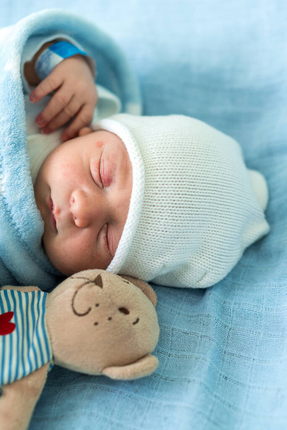 Close-up of Newborn Baby Face Portrait Early Days Sleeping With Tady Bear On Blue Background. Child At Start Minutes Of Life on Hat. Infants, Childbirth, First Moments Of Borning, Beginning Concept. - 写真・画像