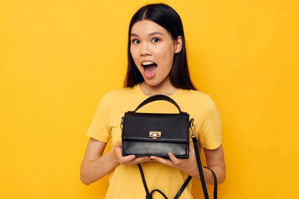 Portrait Asian beautiful young woman with a black womans bag in her hands yellow background unaltered - Valokuva, kuva