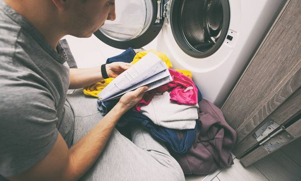 Hombre tratando de cargar la lavadora secadora con ropa, pero no sabe cómo combinar los tipos y colores de la ropa por lo que está leyendo la instrucción - Foto, Imagen