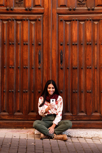 beautiful young indian woman sat over a wooden church door using cellphone, vertical copy space - Foto, Imagen