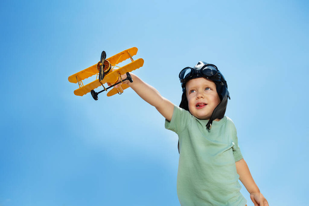 Portrait of the cute boy with airplane toy over blue sky in aviator hat and glasses - Photo, image