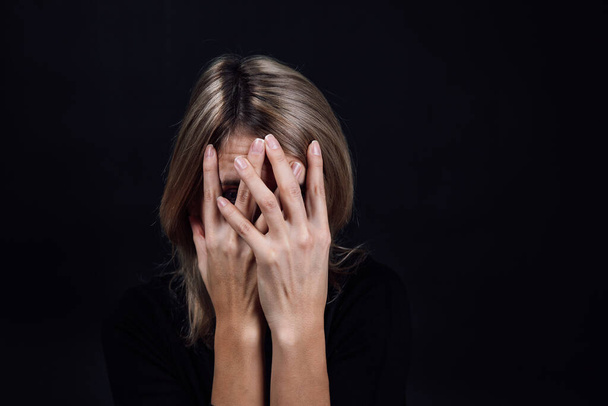 Unhappy young woman in black blouse with hands covering face, eyes looking at camera through fingers on black background. Physical and psychological abuse. Gaslighting - 写真・画像