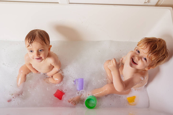 Two siblings play in the bath at home together with toys and bubbles laughing looking to camera top view - Фото, изображение