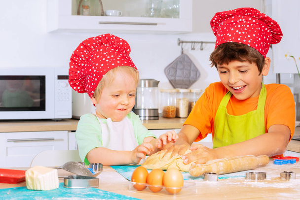 Két fiú tésztát morzsol, séf piros kalapot viselő kezekkel, és mosolyogva ül az asztalnál a konyhában. - Fotó, kép