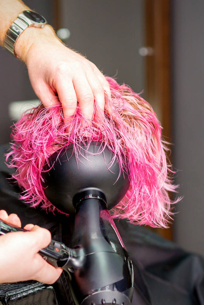 Стилист волос делает прическу с помощью фена, дующего на мокрые традиционные розовые волосы в салоне красоты - Фото, изображение