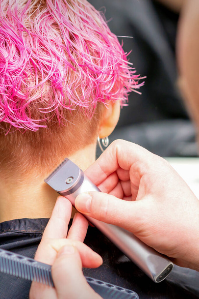 Парикмахер бреет затылок и шею электрическим триммером молодой кавказки с короткими розовыми волосами в салоне красоты - Фото, изображение