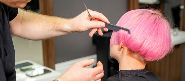Τα χέρια του κομμωτή χτένισμα μαλλιά κάνοντας σύντομο ροζ χτένισμα για μια νεαρή καυκάσια γυναίκα σε ένα σαλόνι ομορφιάς - Φωτογραφία, εικόνα