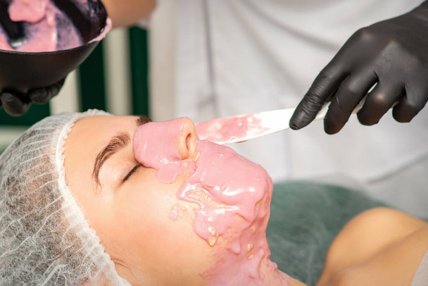 O cosmetologista aplicando uma máscara de alginato no rosto de uma jovem mulher em um salão de beleza - Foto, Imagem