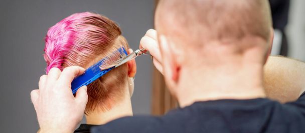 Una mujer con un nuevo corte de pelo. Peluquero masculino cortando pelo corto rosa con tijeras en una peluquería - Foto, Imagen