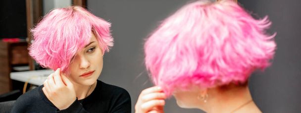 Schöne junge kaukasische Frau, die ihre kurzen rosafarbenen Haare im Spiegel betrachtet - Foto, Bild