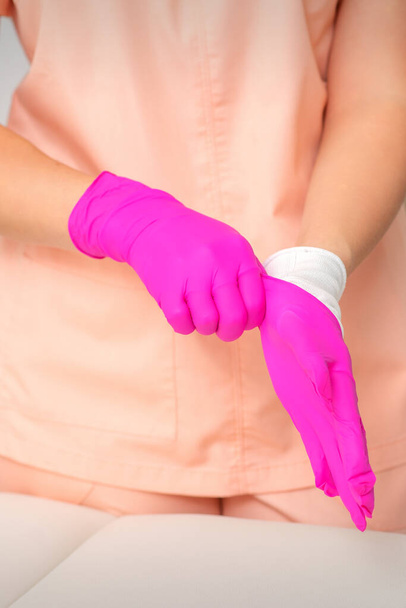 Käsi kosmetologi laittaa steriilit vaaleanpunaiset käsineet valmistautuu vastaanottamaan asiakkaita sisätiloissa - Valokuva, kuva