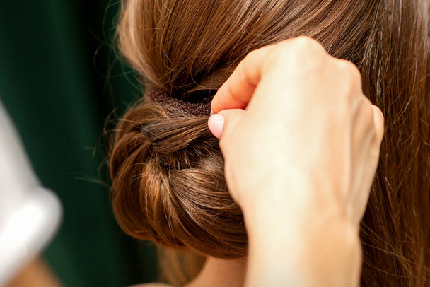 Hände des Friseurs machen Französisch Twist Frisur einer unkenntlichen jungen brünetten Frau in einem Schönheitssalon, Rückansicht, Nahaufnahme - Foto, Bild