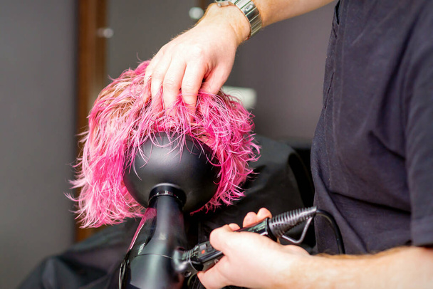 Στυλίστρια μαλλιών κάνει χτένισμα χρησιμοποιώντας στεγνωτήρα μαλλιών φυσώντας σε βρεγμένα έθιμο ροζ μαλλιά σε ένα σαλόνι ομορφιάς - Φωτογραφία, εικόνα