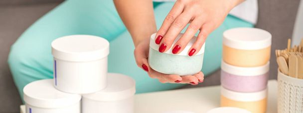 Руки женщины открывают банку крема для тела поверх стола со множеством банок косметики - Фото, изображение