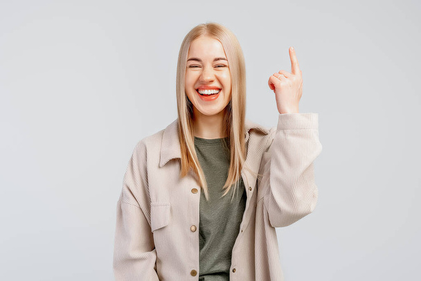 Ευτυχισμένο κομψό ξανθό κορίτσι σε μοντέρνο πουκάμισο δείχνοντας το δάχτυλο προς τα πάνω και χαμογελώντας, καλώντας να εγγραφείτε, δείχνει το λογότυπο της εταιρείας, γκρι φόντο. - Φωτογραφία, εικόνα