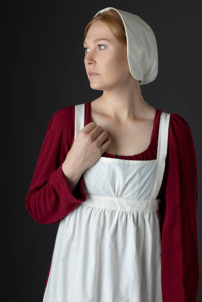 Una sirvienta de la Regencia con un vestido de lino rojo con un delantal y una gorra contra un fondo de estudio - Foto, imagen