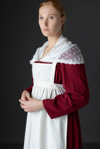 A Regency maid or working class woman wearing a red linen dress with a modest shawl and apron against a studio backdrop - Photo, Image