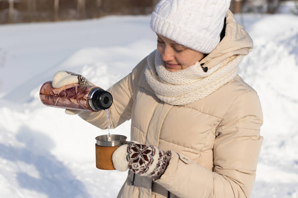 Una hermosa joven con chaqueta y manoplas vierte una bebida caliente en una taza de un termo en la naturaleza en un día soleado de invierno. Enfoque selectivo. Retrato. Primer plano - Foto, imagen