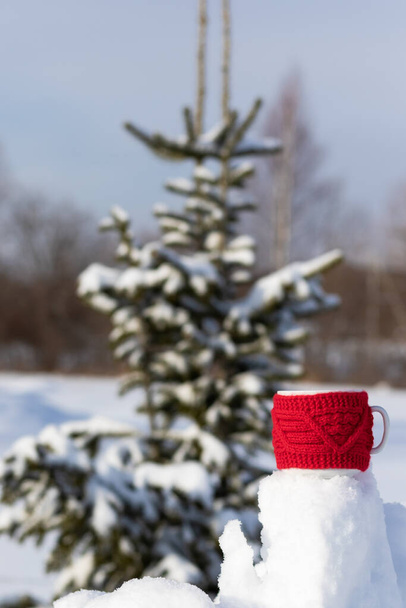 A red mug with a hot drink on a snowdrift on a frosty sunny winter day in the forest against the backdrop of snow-covered fir trees. Landscape. Selective focus. Close-up - Photo, Image
