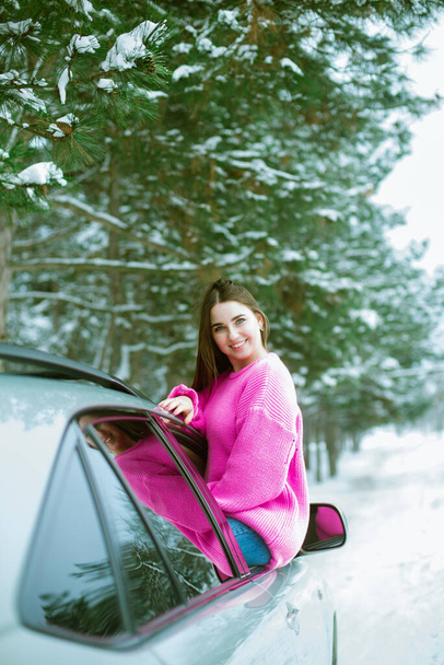 Young cute girl in a pink sweater laughs and looks out of the car window on a winter road in the snow  - Fotoğraf, Görsel