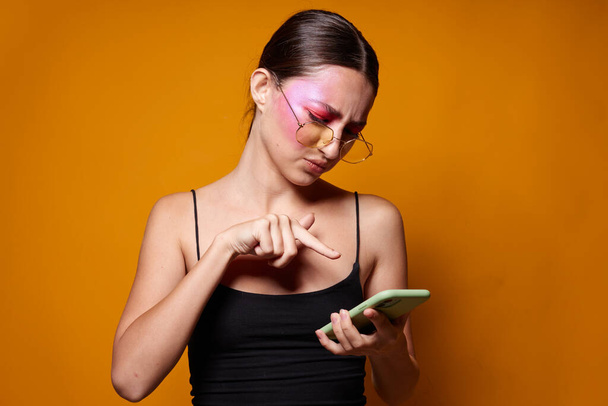 Sexy brunette woman with phone pink face makeup posing attractive look glasses yellow background unaltered - Photo, Image